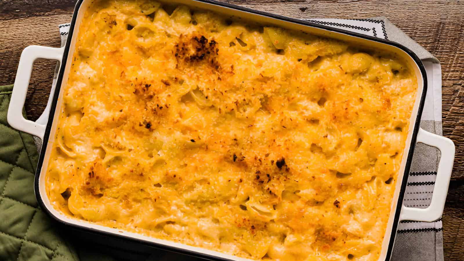 A rectangular dish filled with baked macaroni and cheese, topped with a golden-brown crust, on a wooden surface next to a green oven mitt.