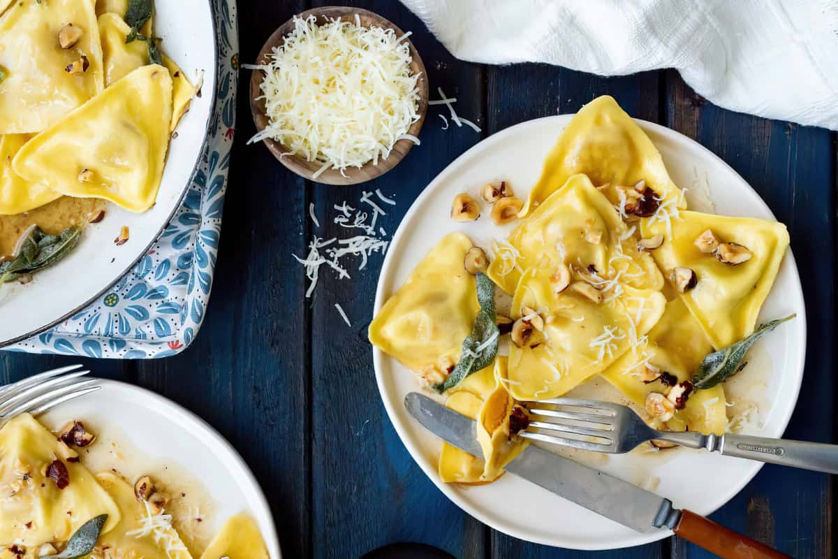 A plate of ravioli garnished with cheese, walnuts, and sage, accompanied by a bowl of grated cheese.