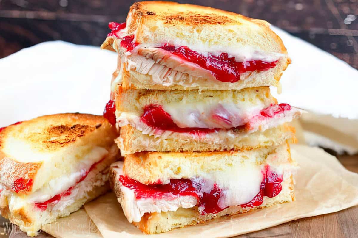 A stack of grilled turkey and cheese sandwiches with cranberry sauce oozing out, served on parchment paper. The sandwiches have crispy, golden-brown bread and melted cheese. A white cloth is in the background.