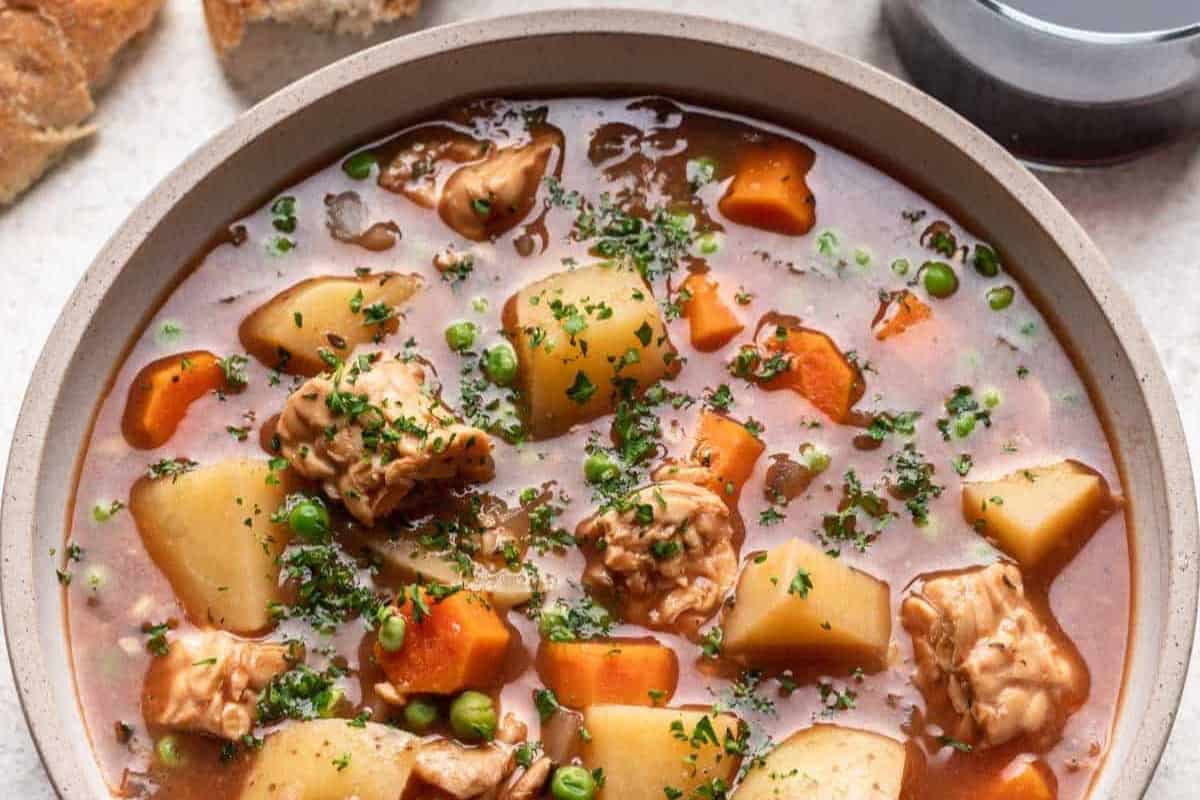 A bowl of hearty stew filled with chunks of meat, potatoes, carrots, and peas in a rich broth. The dish is topped with freshly chopped herbs. Pieces of bread are visible in the background.