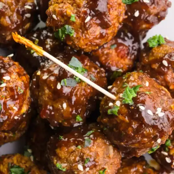 A close-up of glazed meatballs topped with sesame seeds and chopped herbs, with a toothpick in one meatball.