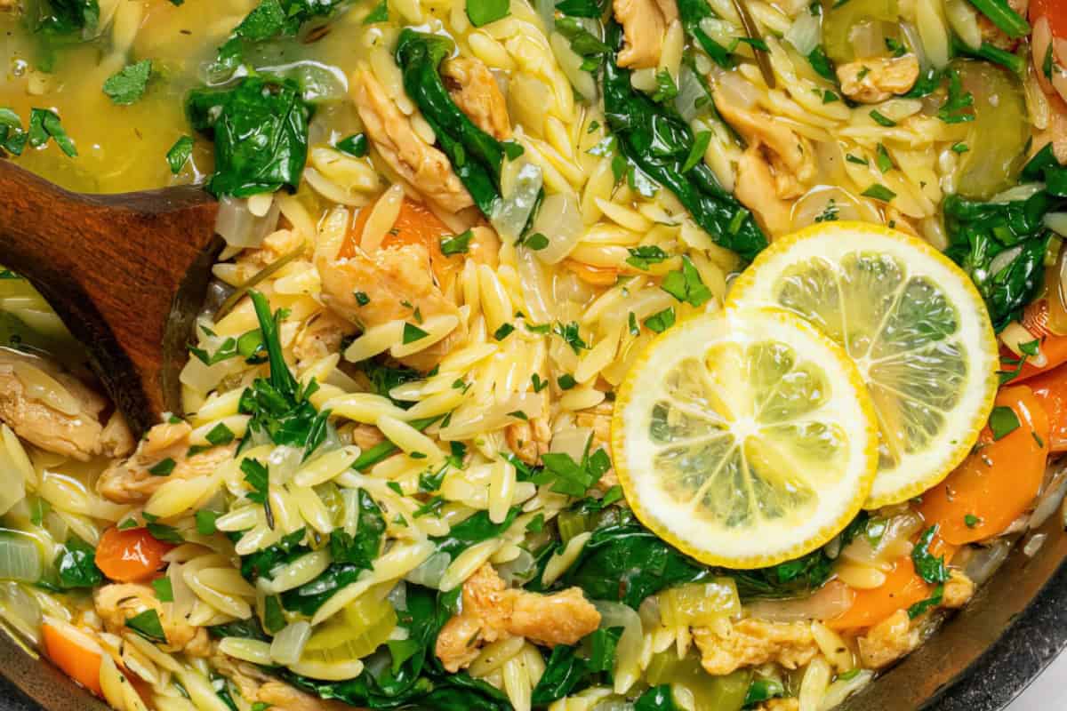 The image shows orzo pasta mixed with spinach, chicken, and various vegetables. It is garnished with two lemon slices and sprinkled with herbs.