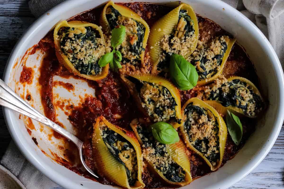 A white baking dish contains stuffed pasta shells topped with breadcrumbs and basil, served over tomato sauce with a spoon on the side.