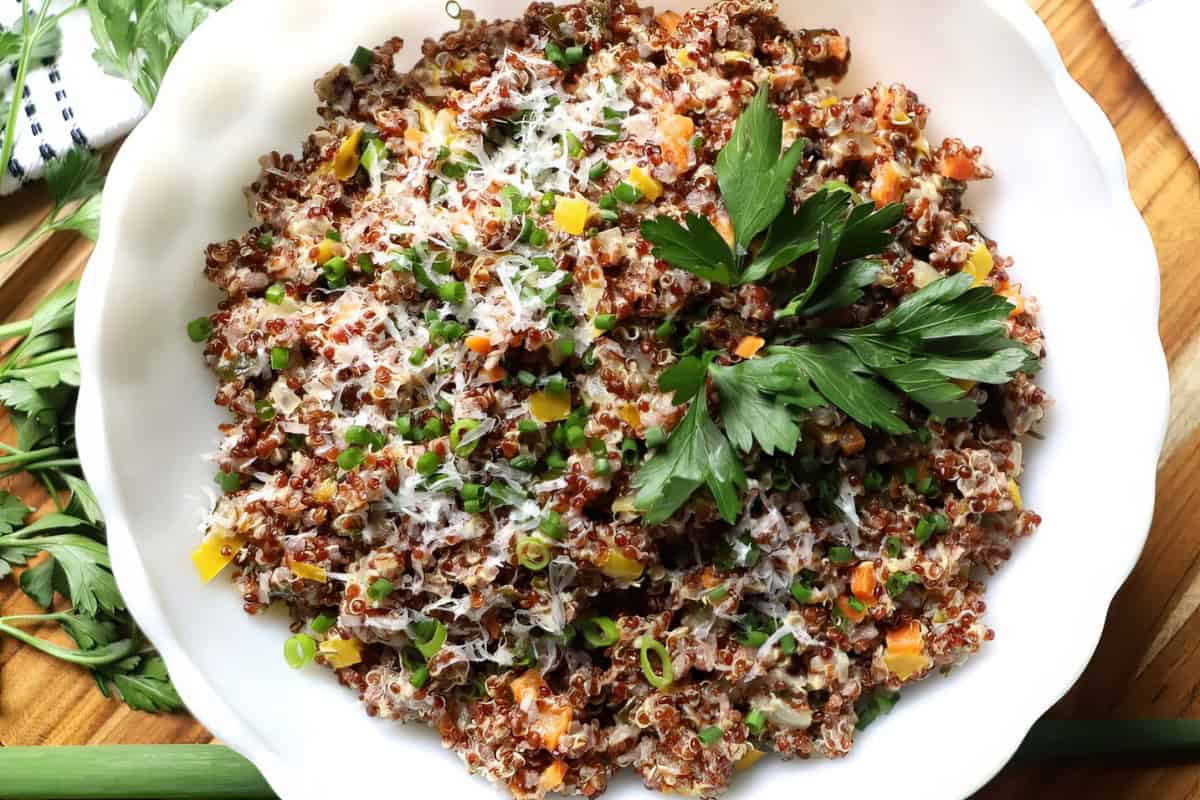 A white bowl filled with a quinoa salad containing a mix of red quinoa, peas, carrots, and yellow bell peppers, topped with grated cheese and garnished with fresh parsley. It's set on a wooden table with green herbs nearby.