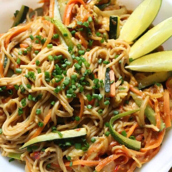 A bowl of noodles with zucchini, carrots, chopped chives, and lime wedges.