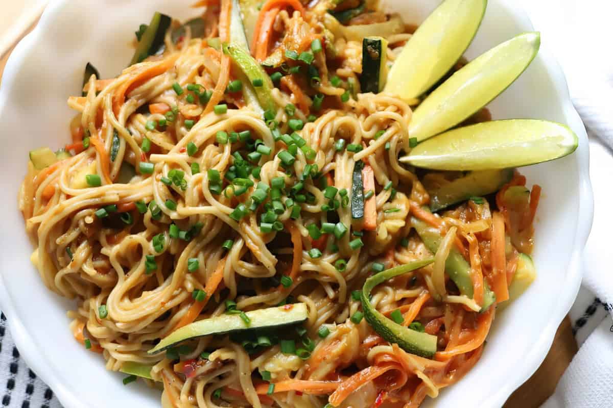 A bowl of noodles with zucchini, carrots, chopped chives, and lime wedges.
