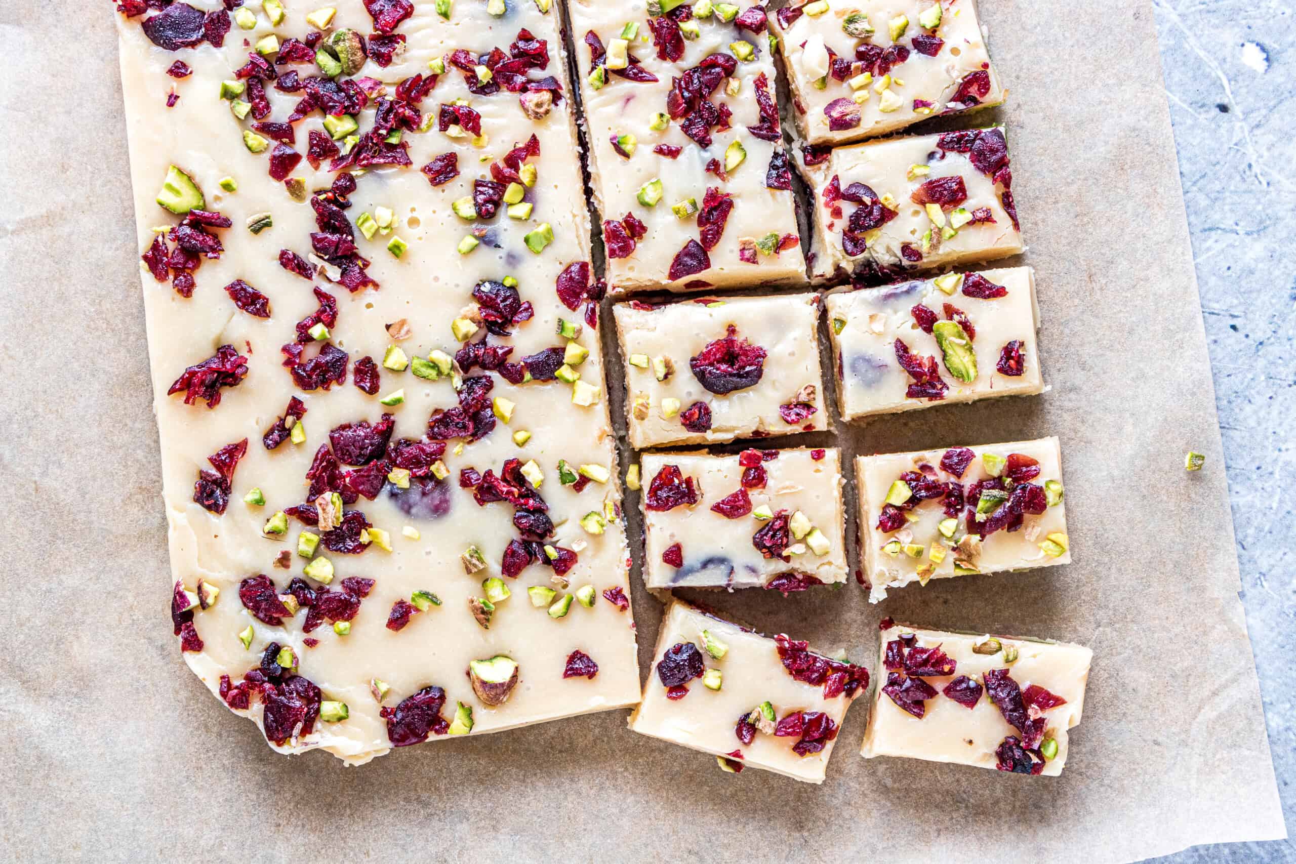Sliced white fudge topped with cranberries and pistachios on parchment paper.