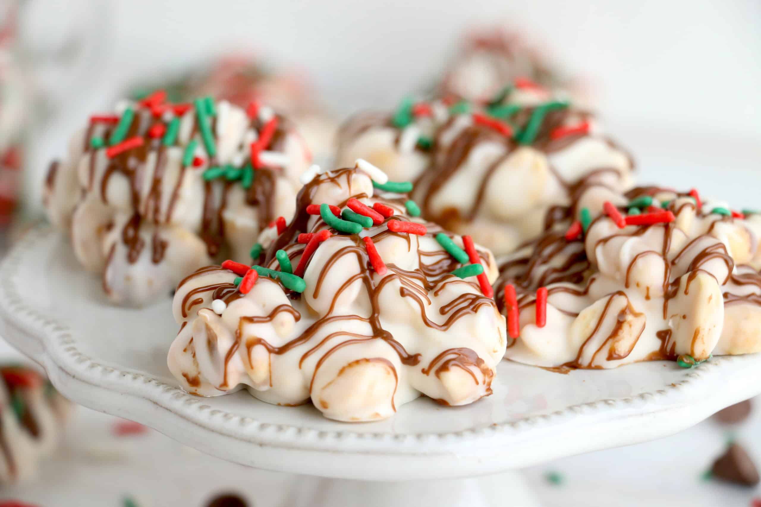 A white serving plate holds clusters of white chocolate, drizzled with milk chocolate and topped with red and green sprinkles.