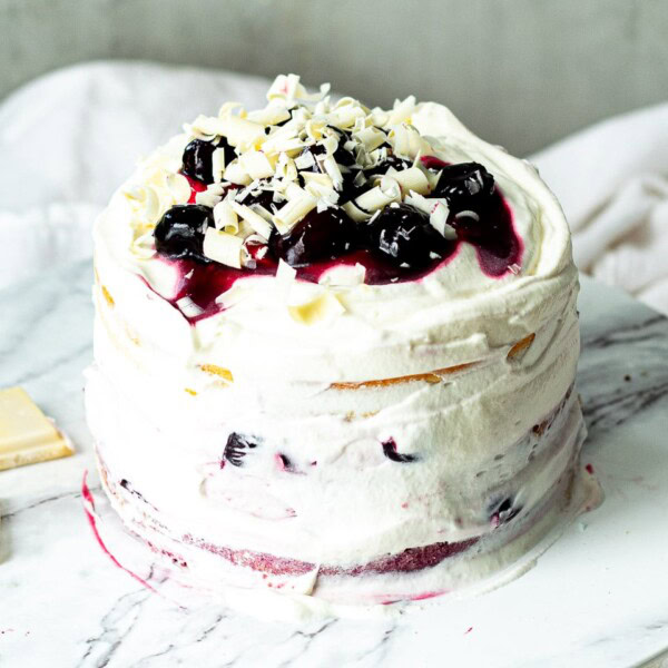 Layered cake with white frosting, dark cherries, cherry sauce, and white chocolate shavings on a marble surface.