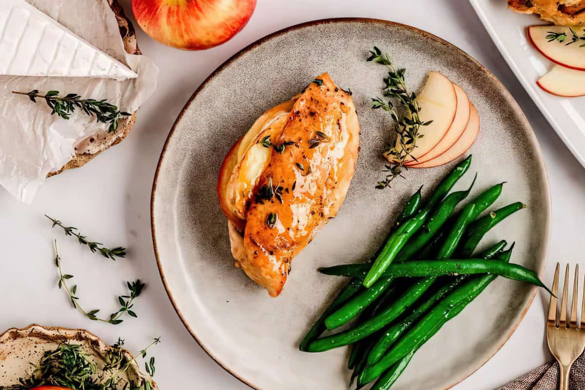 A plate with a chicken breast topped with herbs, fresh green beans, and apple slices. Nearby are a wedge of brie cheese, a sprig of thyme, and a whole apple, all set on a light surface with a fork beside the plate.