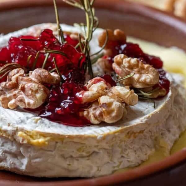 A round baked brie topped with walnuts, red jam, and rosemary on a brown plate with melted cheese pooling at the edges.