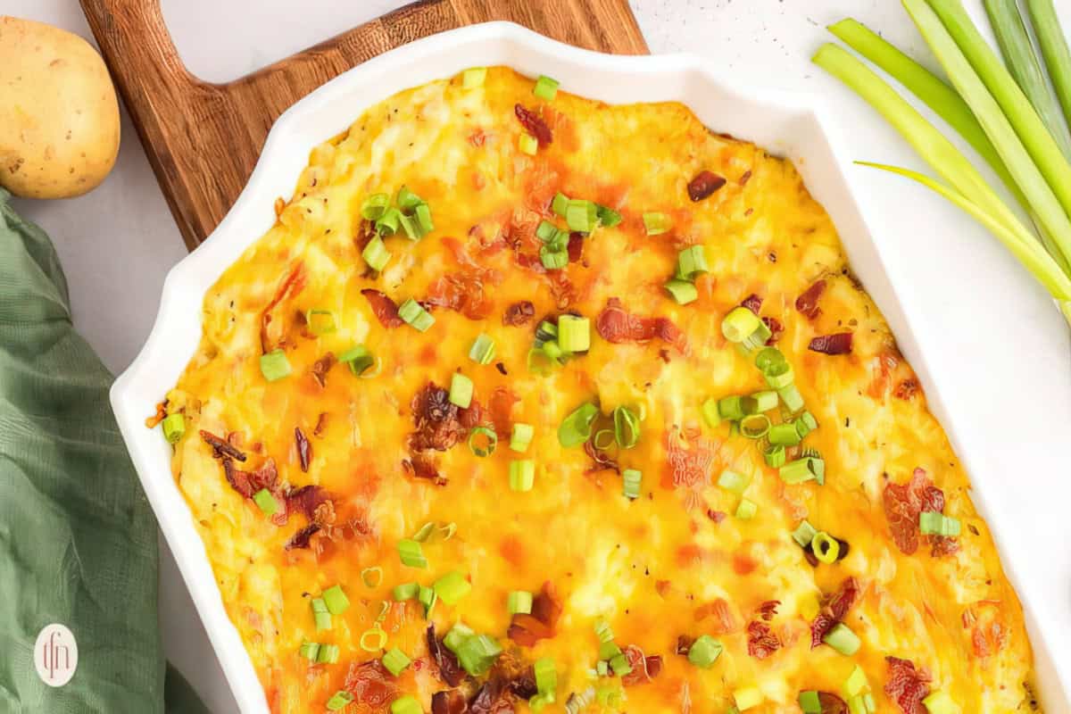 A cheesy baked dish garnished with chopped green onions and crispy bits in a white casserole dish. It rests on a wooden board beside a green napkin and fresh scallions.