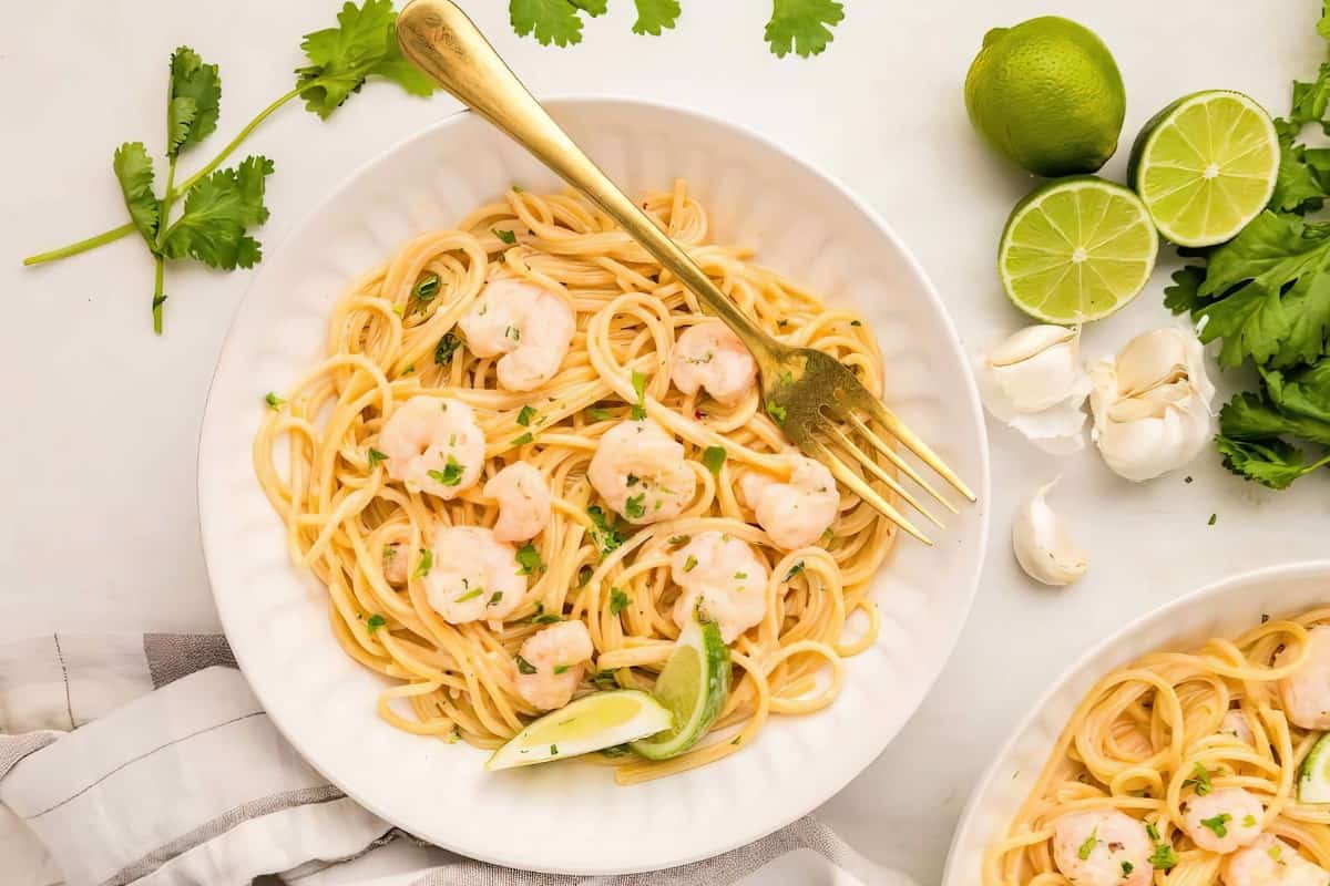 A plate of spaghetti with shrimp, lime wedges, and herb garnish. Nearby are fresh cilantro, lime, and garlic. A gold fork rests on the plate.