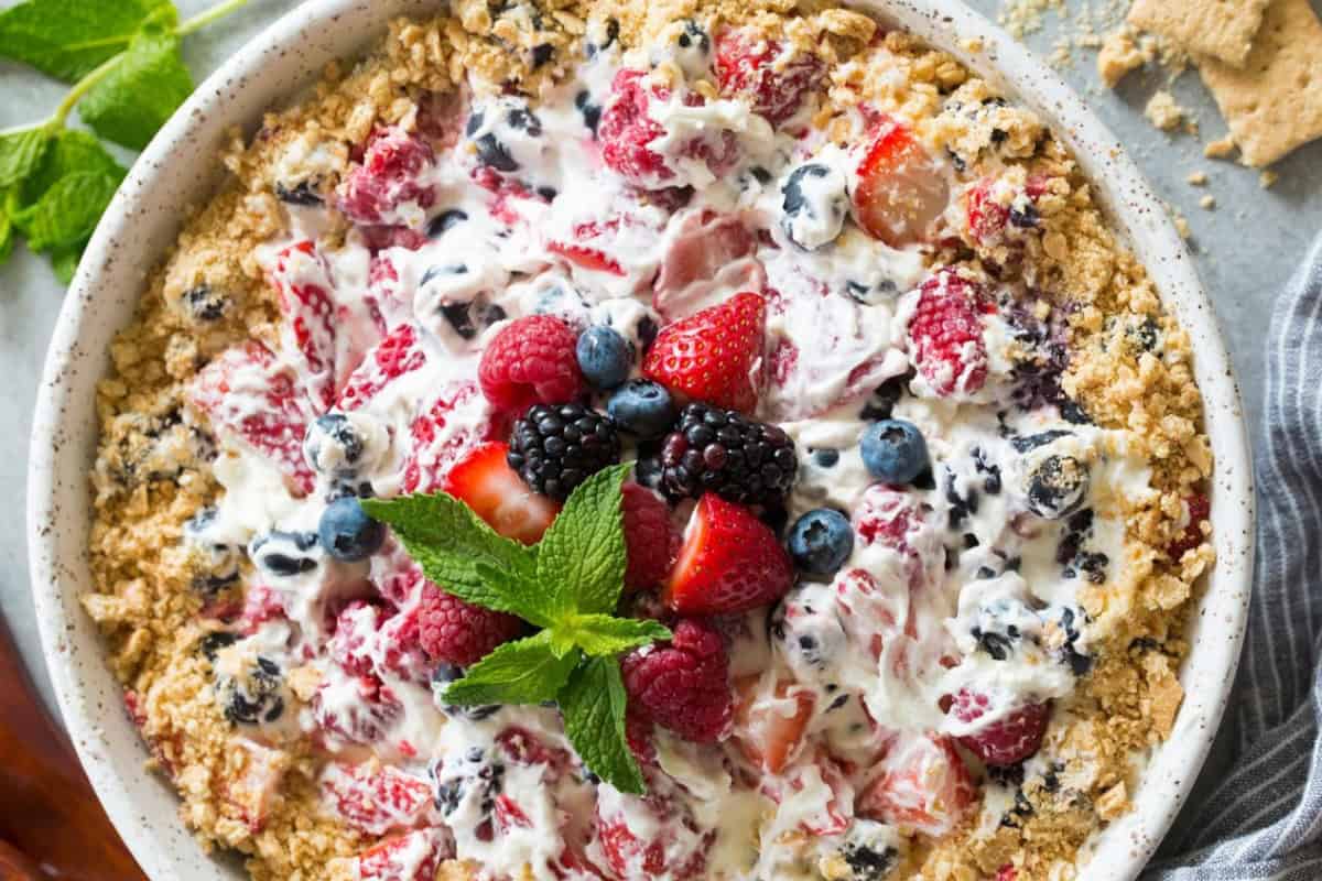 A creamy dessert topped with mixed berries, including strawberries, raspberries, blackberries, and blueberries. Crumbled topping surrounds the dish, which is garnished with fresh mint leaves in the center.