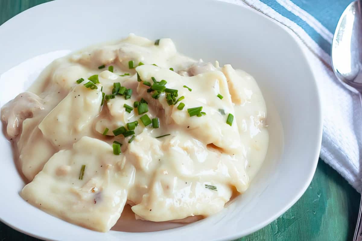 A white plate filled with creamy chicken and dumplings, garnished with chopped chives, sits on a green table. A spoon and white napkin are placed beside the plate.