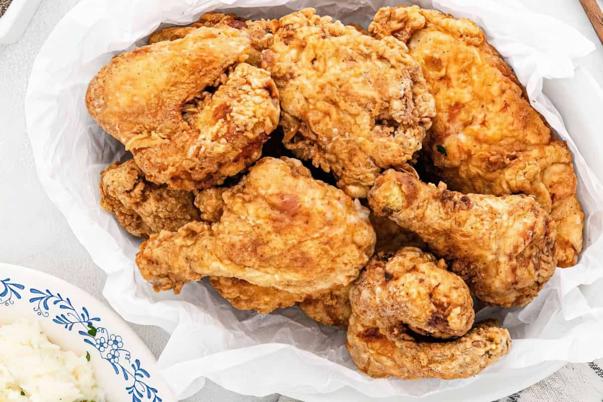 The image features a basket of crispy fried chicken pieces on parchment paper, accompanied by a plate of mashed potatoes.
