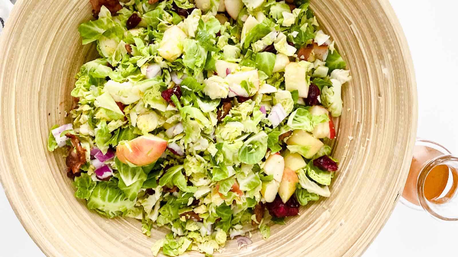 A large wooden bowl contains a fresh chopped salad with Brussels sprouts, apples, cranberries, red onions, and a dressing bottle placed beside the bowl.