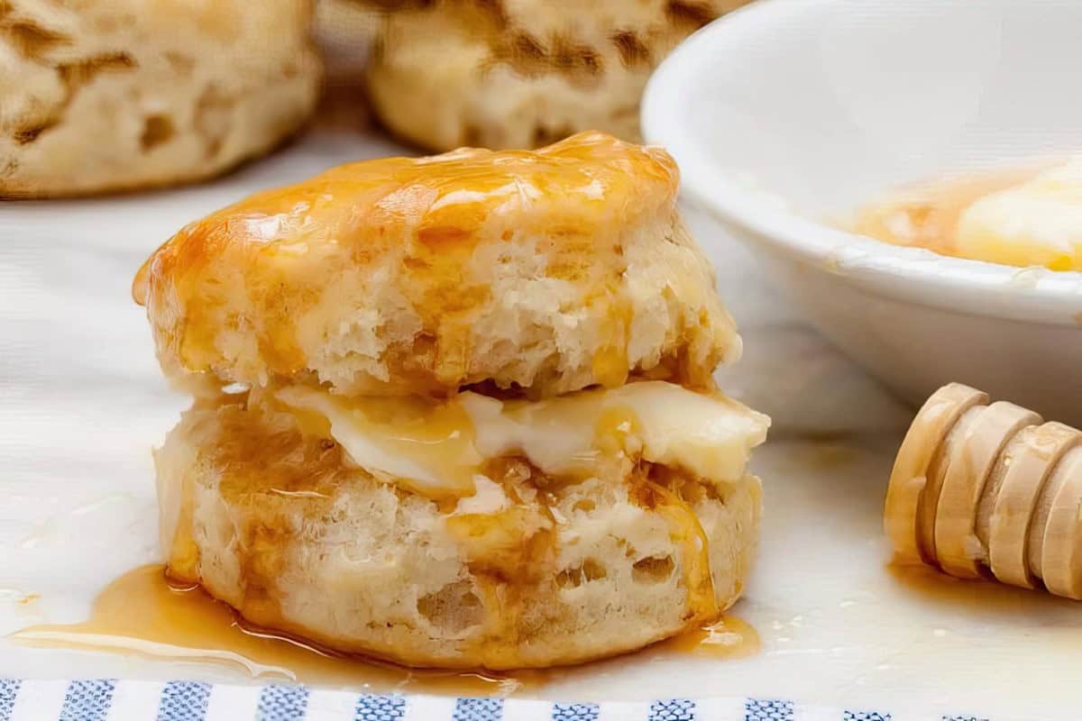 A biscuit split open with butter and honey is on a checkered cloth, accompanied by a partially visible bowl and honey dipper in the background.
