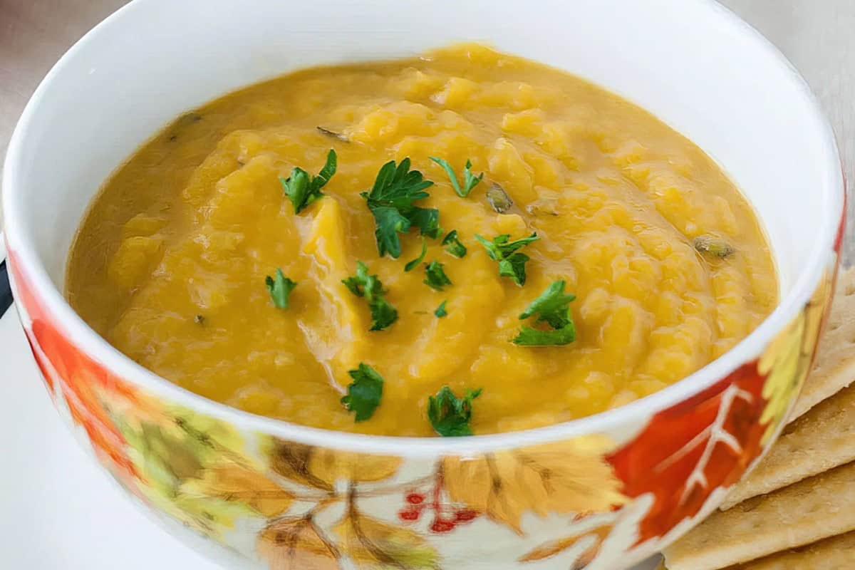 A bowl of creamy pumpkin soup garnished with fresh parsley. The bowl has autumn leaf patterns, and there's a glimpse of crackers on the side.