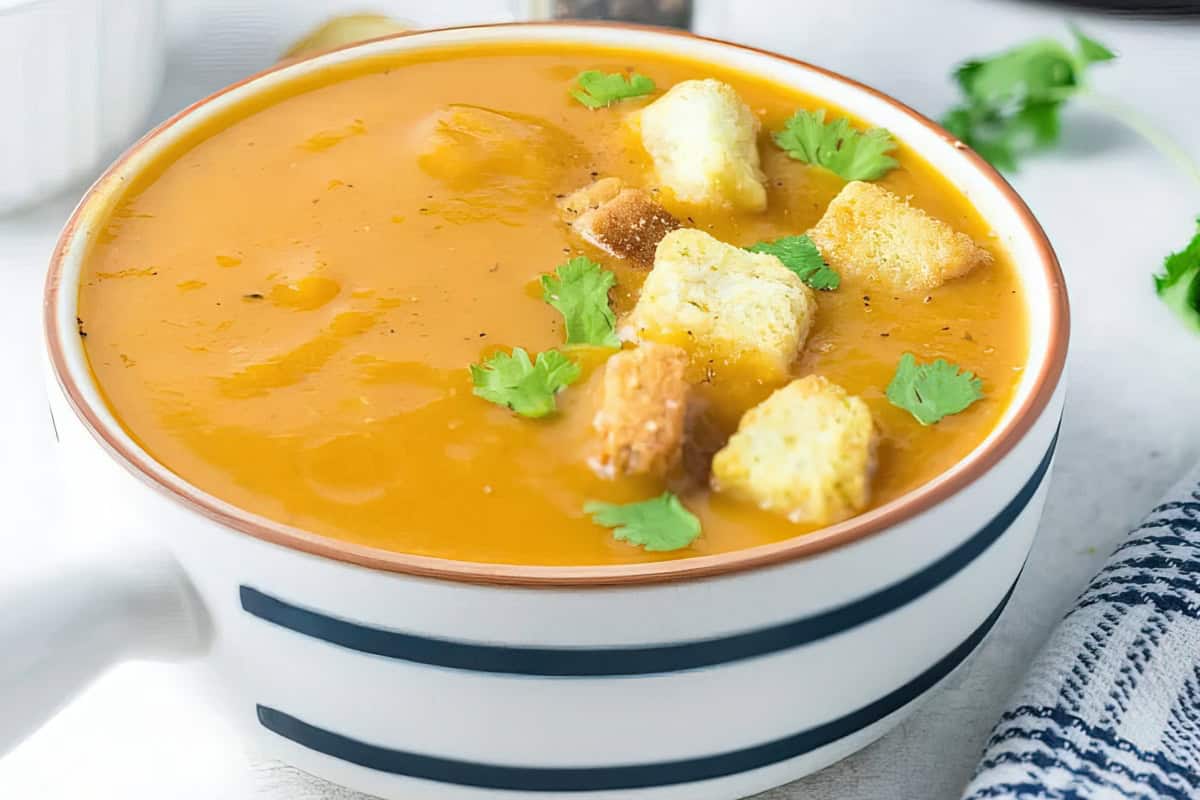A bowl of creamy orange soup topped with croutons and cilantro leaves. The soup is in a white bowl with black stripes, set on a white surface near a striped cloth napkin.