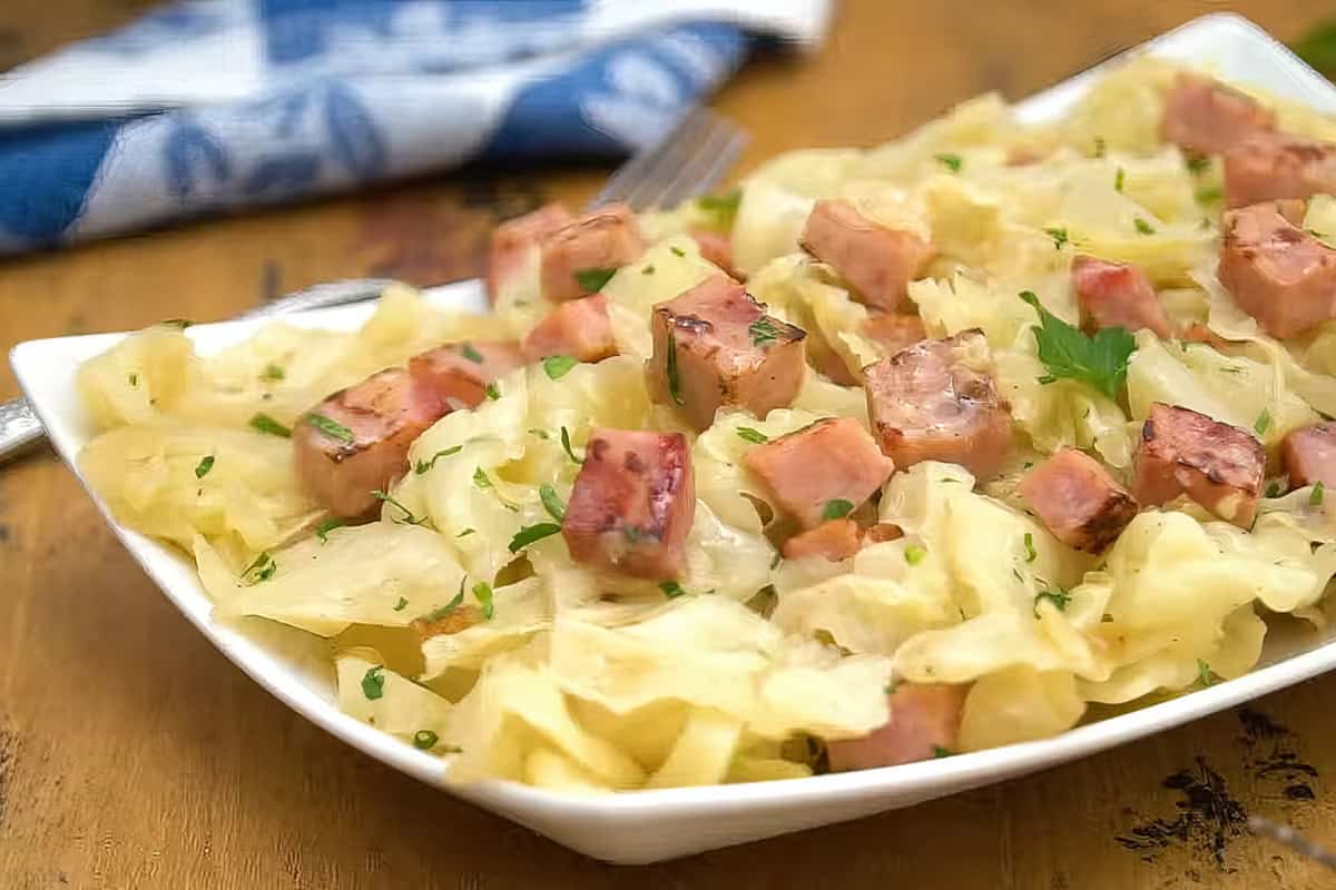 A platter of cooked cabbage and diced ham, garnished with parsley, is served on a white dish. A blue and white napkin is in the background on a wooden table.