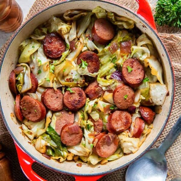 A pot of cooked cabbage and sliced sausage with herbs sits on a table beside a spoon, pepper mills, and fresh parsley.