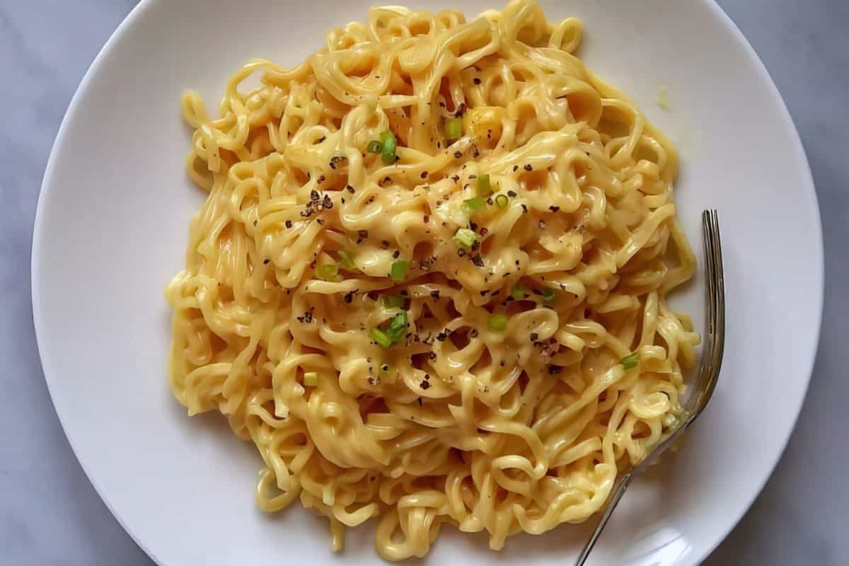 The image shows a plate of creamy cheese noodles topped with chopped green onions and black pepper, alongside a fork.