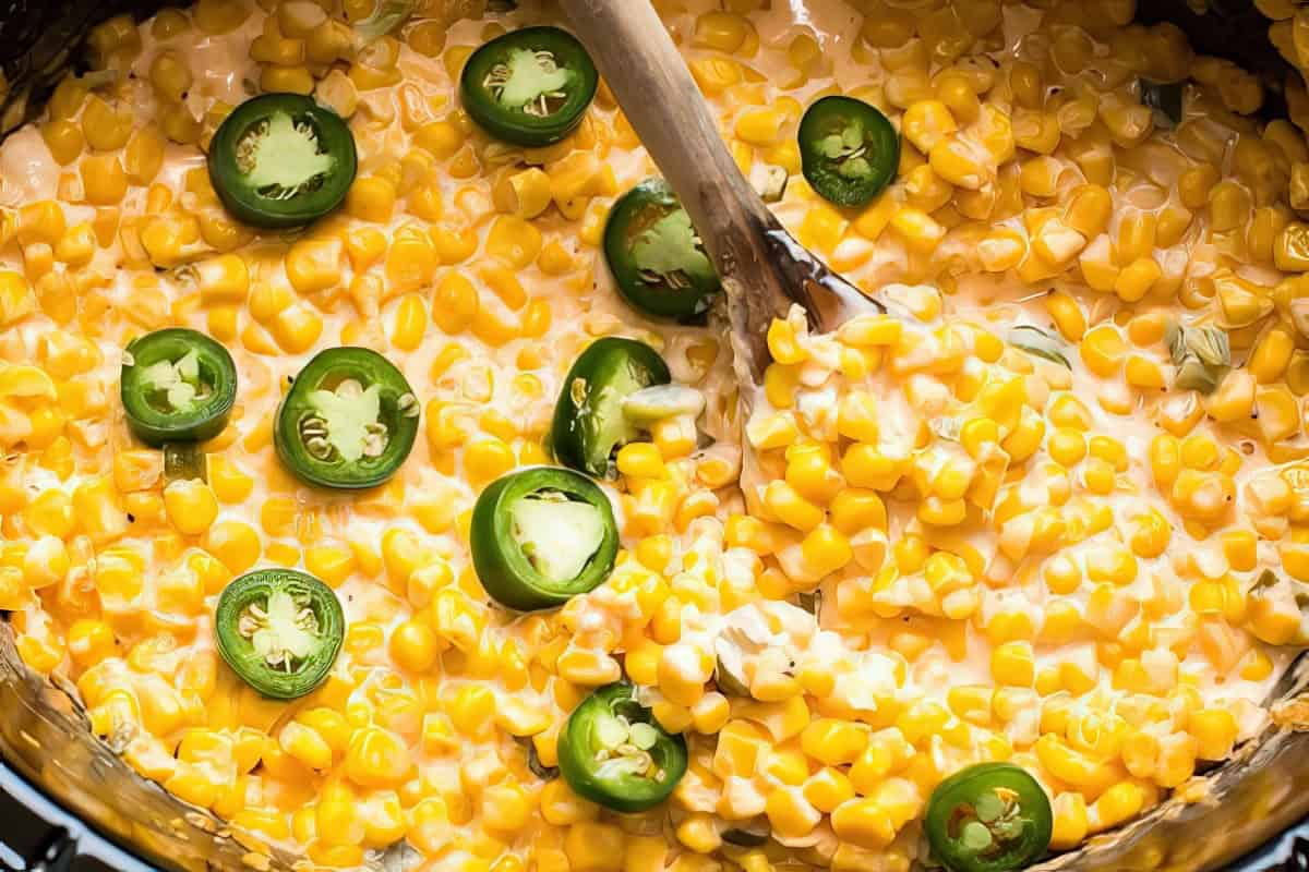 Close-up of creamy corn in a pot, mixed with slices of green jalapeño peppers. A wooden spoon is stirring the mixture.