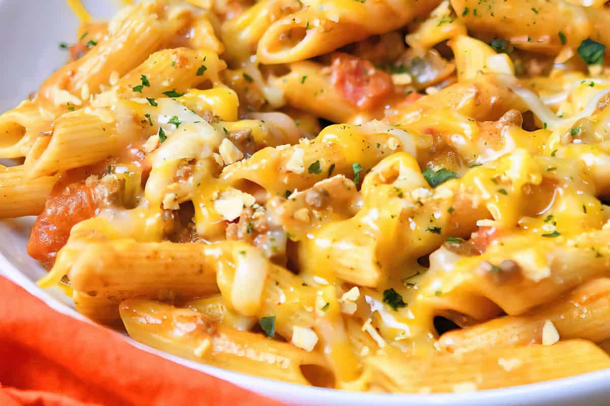 The image shows a close-up of penne pasta mixed with ground meat, covered in melted cheese. There are visible pieces of tomato and a sprinkle of herbs on top.