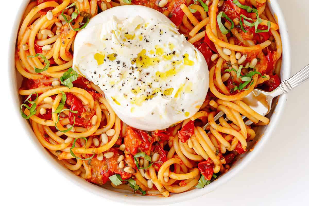 The image depicts a bowl of spaghetti topped with burrata cheese, pine nuts, basil, and a drizzle of olive oil.
