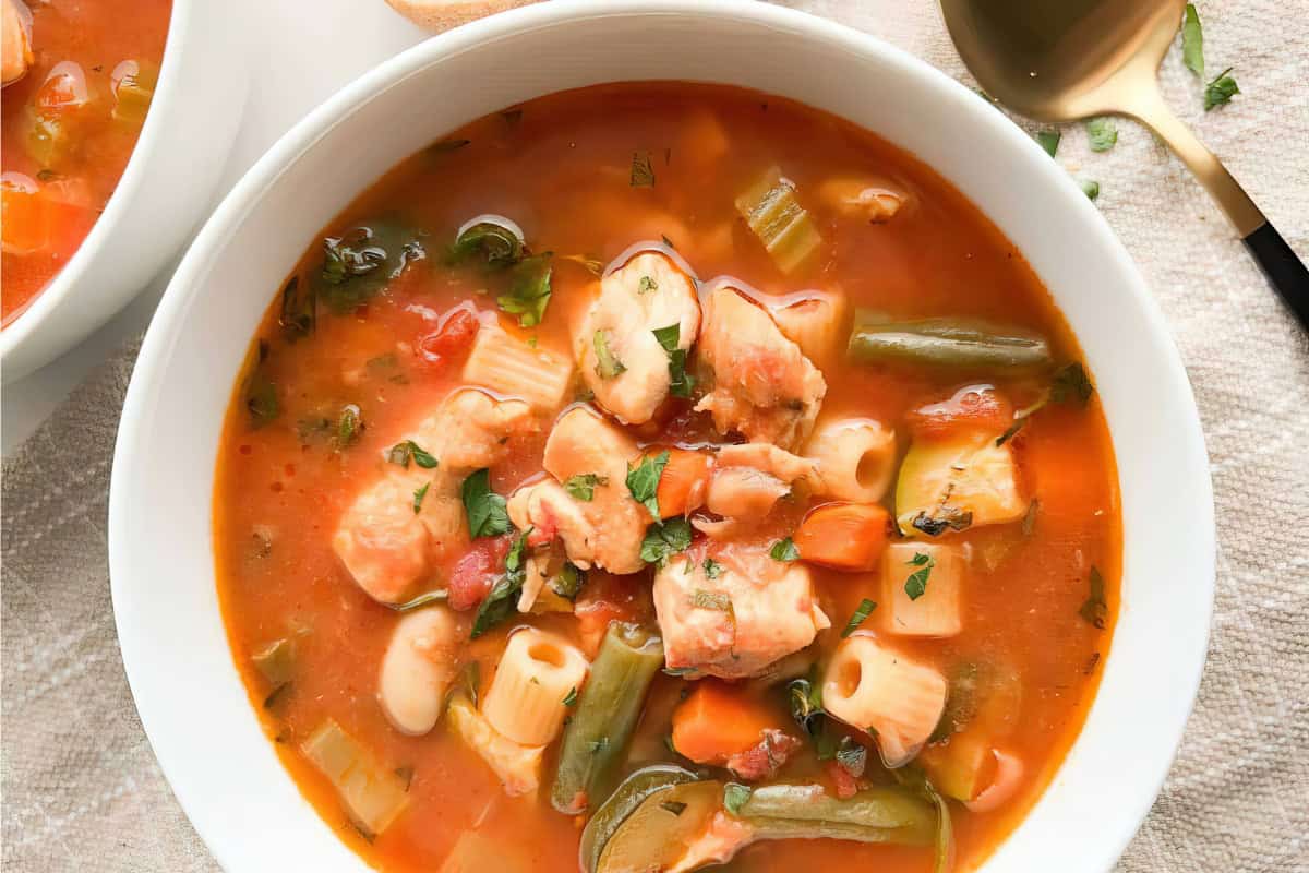 A bowl of soup containing pasta, vegetables, and pieces of chicken in a tomato broth.