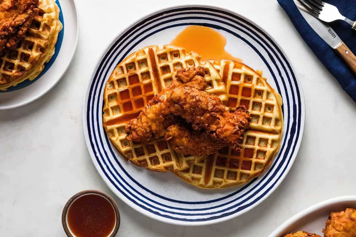 A plate with two waffles topped with crispy fried chicken, drizzled with syrup. Accompanied by a fork and knife on a napkin, and a small dish of syrup on the side.