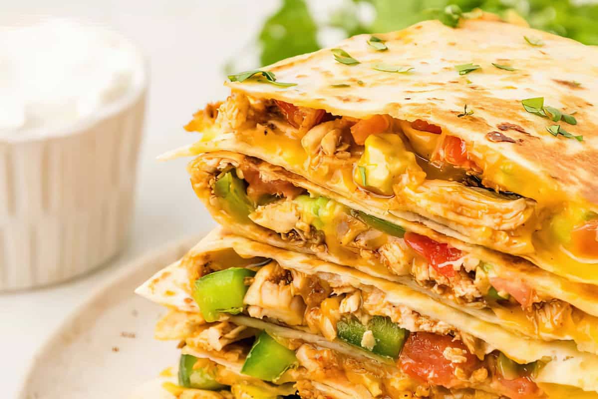 A stack of quesadillas filled with melted cheese, bell peppers, and tomatoes on a white plate. A cup of sour cream is in the background.