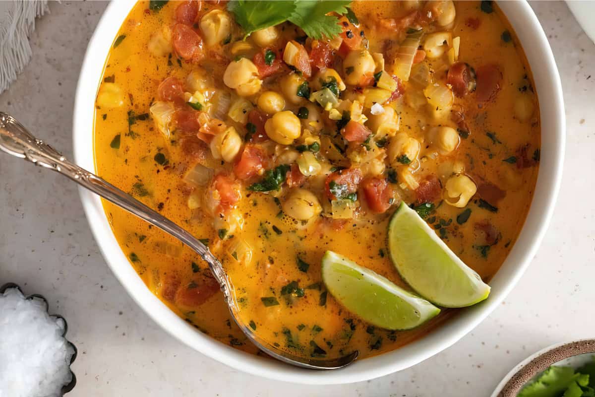 The image depicts a bowl containing creamy chickpea curry, garnished with lime wedges and cilantro. A silver spoon rests in the bowl.