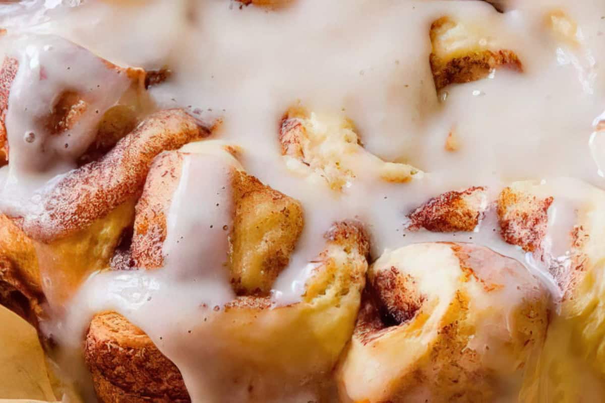 Close-up of freshly baked cinnamon rolls topped with creamy vanilla icing, glistening under warm lighting. The rolls are soft and golden brown, showcasing swirls of cinnamon and sugar.