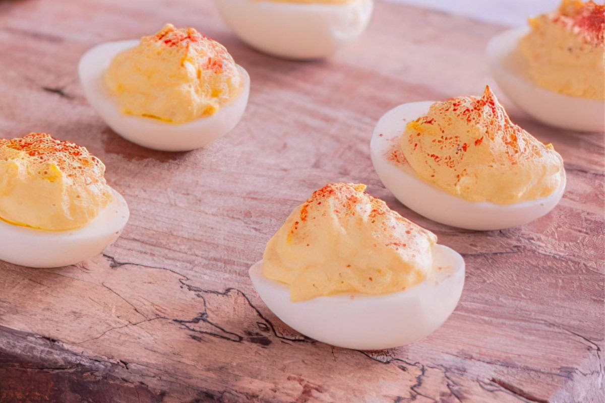 Deviled eggs with a creamy yolk filling, sprinkled with paprika, are arranged on a wooden board.