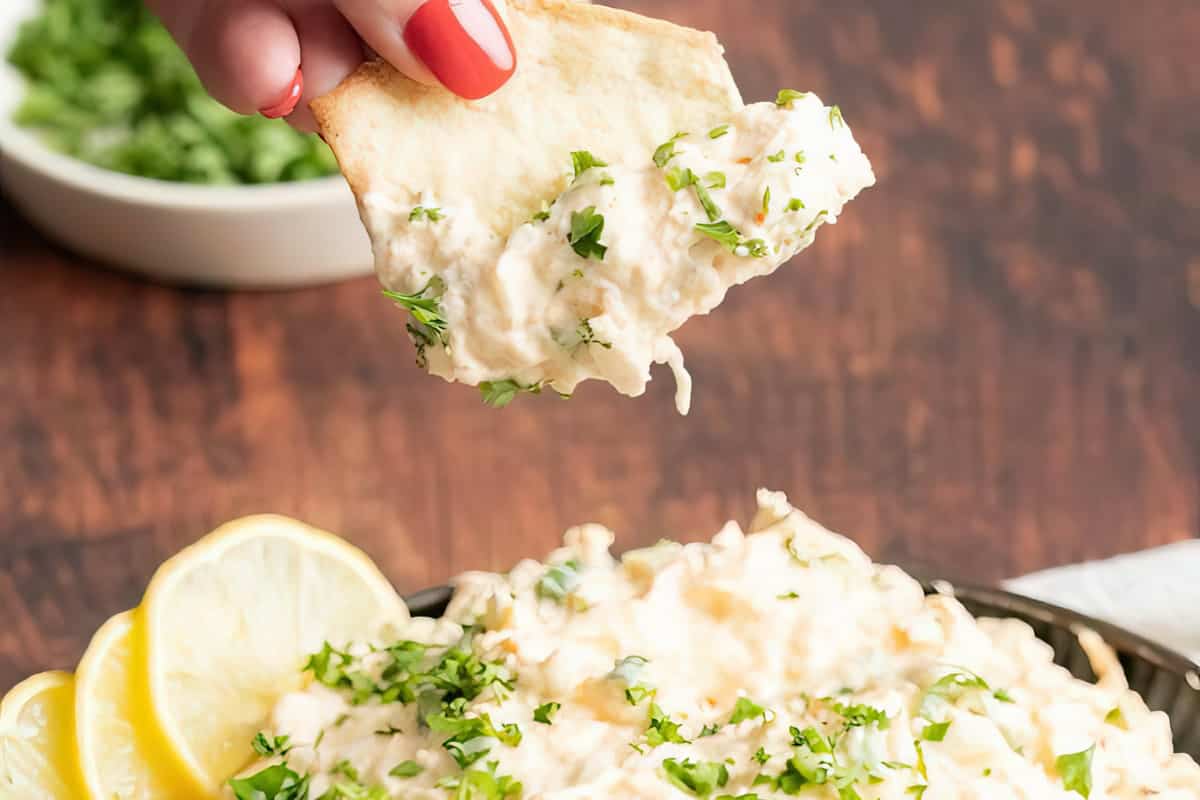 A hand holds a pita chip topped with creamy dip and garnished with chopped herbs, with lemon slices in the background.