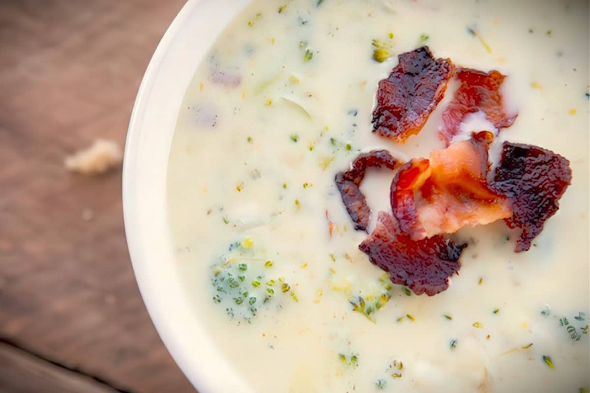 A bowl of creamy soup topped with crispy bacon and broccoli pieces, placed on a rustic wooden surface.