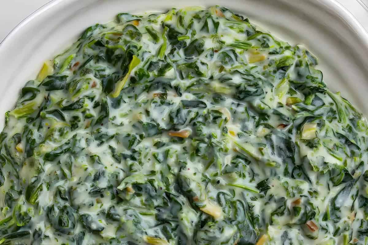 A close-up of a dish of creamed spinach in a white bowl. The spinach is mixed with a creamy sauce, creating a smooth and textured appearance with visible green leaves throughout.