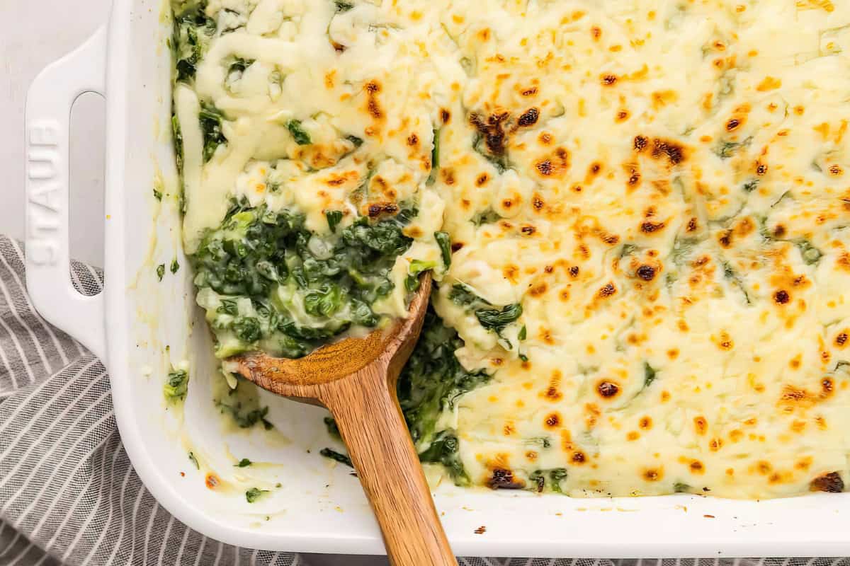 A white baking dish containing a creamy spinach lasagna, partially served with a wooden spoon.