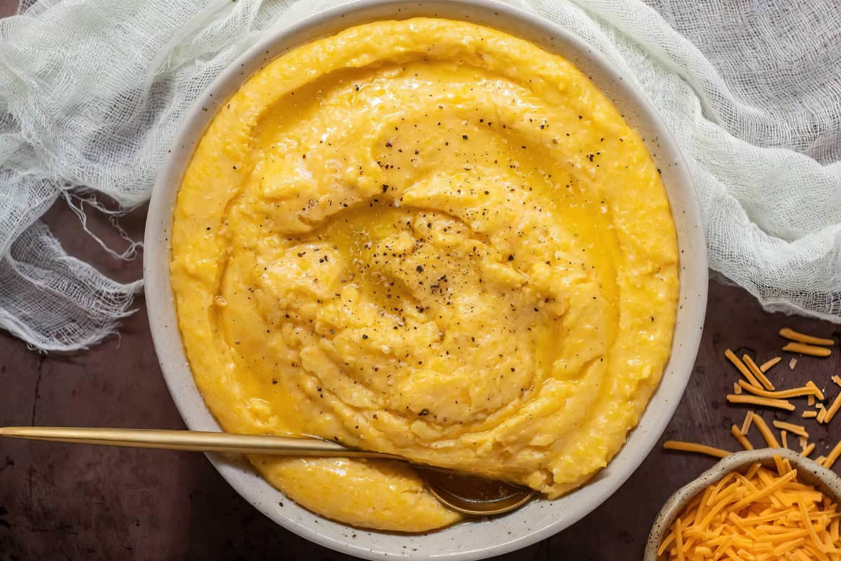 A bowl of yellow grits with black pepper on top, a gold spoon inside, and shredded cheese on the side.