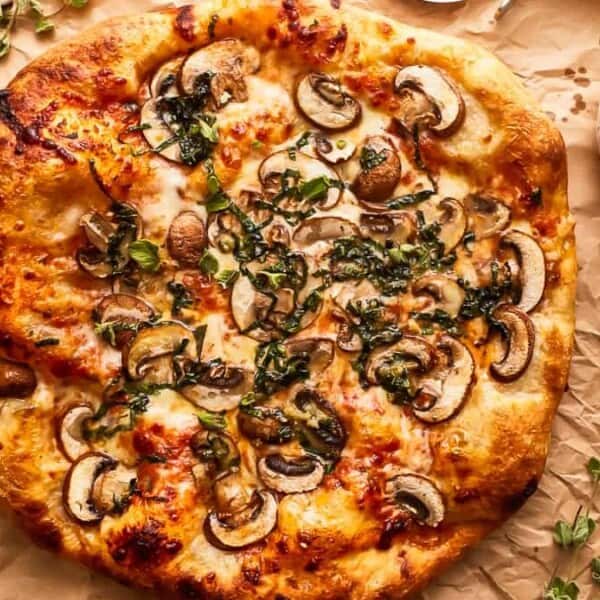 A pizza topped with mushrooms, cheese, and herbs on crumpled parchment paper beside a bowl of olive oil and a brush.