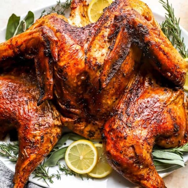 A roasted spatchcock chicken on a white platter, garnished with lemon slices and fresh herbs.