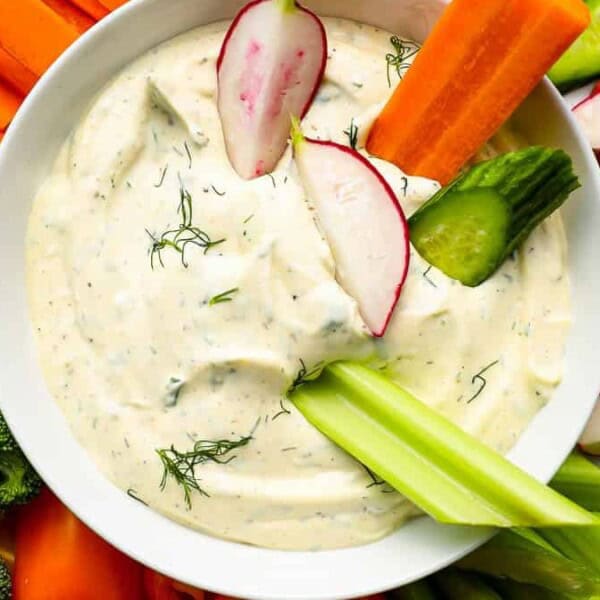 A bowl of creamy dip topped with dill and sliced vegetables, surrounded by broccoli, carrots, celery, cucumber, and radishes.