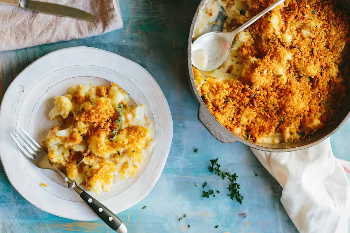 A plate of macaroni and cheese topped with breadcrumbs sits next to a cast iron skillet containing additional servings of macaroni and cheese.