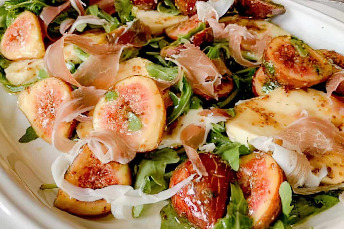 A fresh salad is arranged on a white plate, featuring sliced figs, prosciutto, arugula, and slices of mozzarella cheese, drizzled with balsamic glaze. The combination of colors and textures creates an appetizing arrangement.