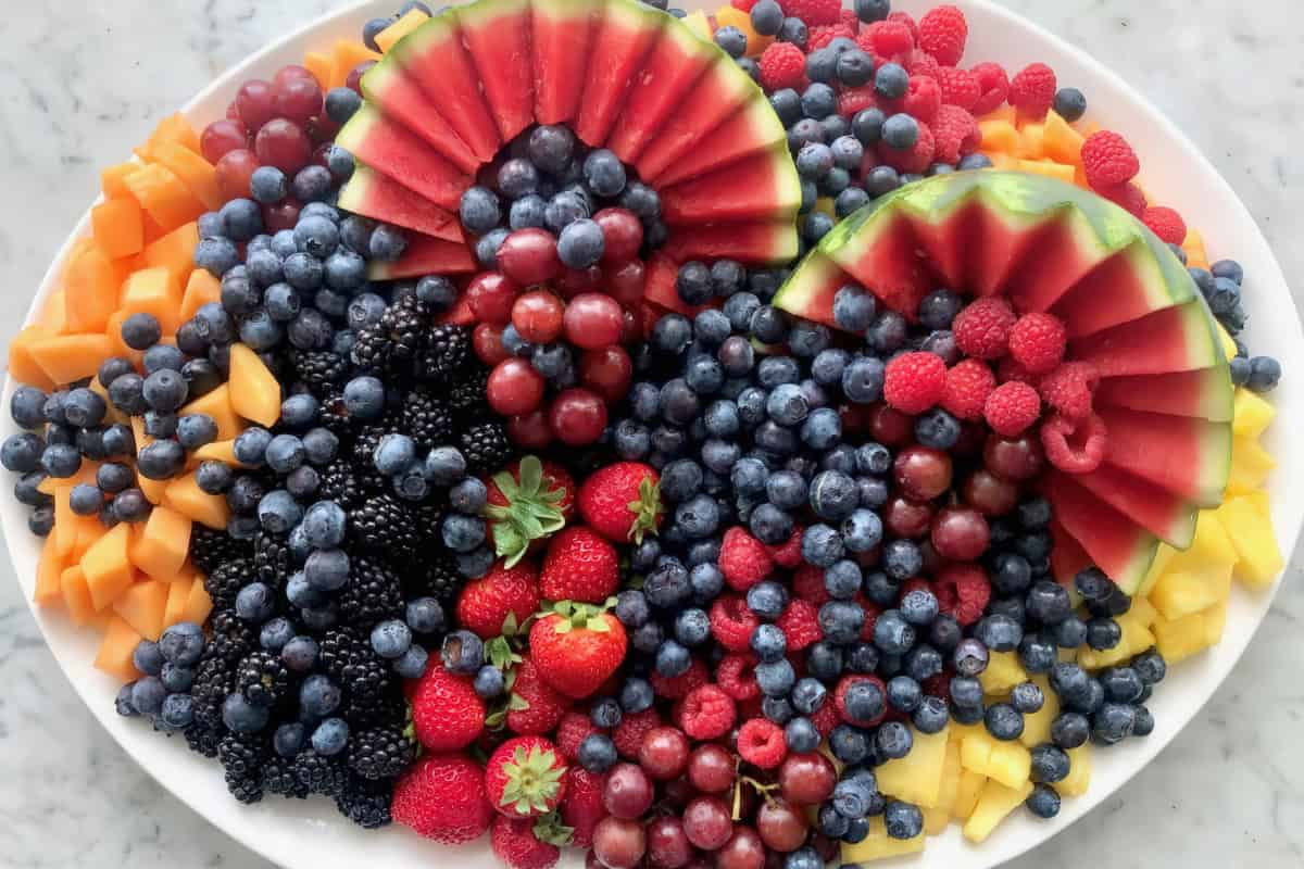 A platter filled with assorted fruits: watermelon slices, blueberries, strawberries, blackberries, raspberries, grapes, mango cubes, and papaya cubes, arranged on a white surface.