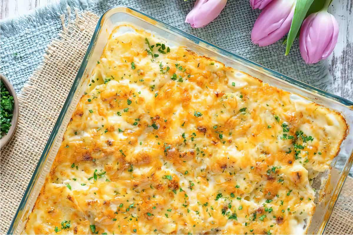 A glass dish with a baked casserole topped with melted cheese and herbs, placed on a burlap mat. Pink tulips are nearby.