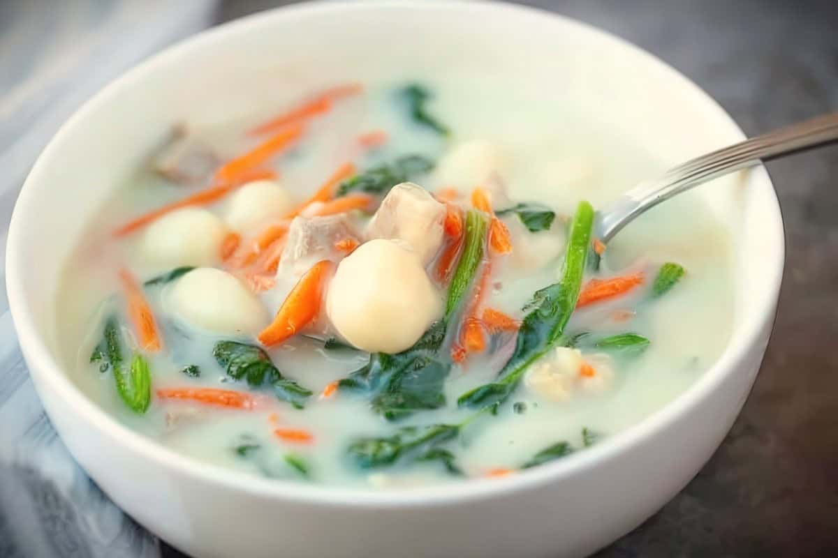 A white bowl filled with creamy soup contains vegetables, leafy greens, carrots, and round dumplings, with pieces of meat. A spoon rests in the bowl, with a striped cloth partially visible beside it. The website URL is visible at the bottom.