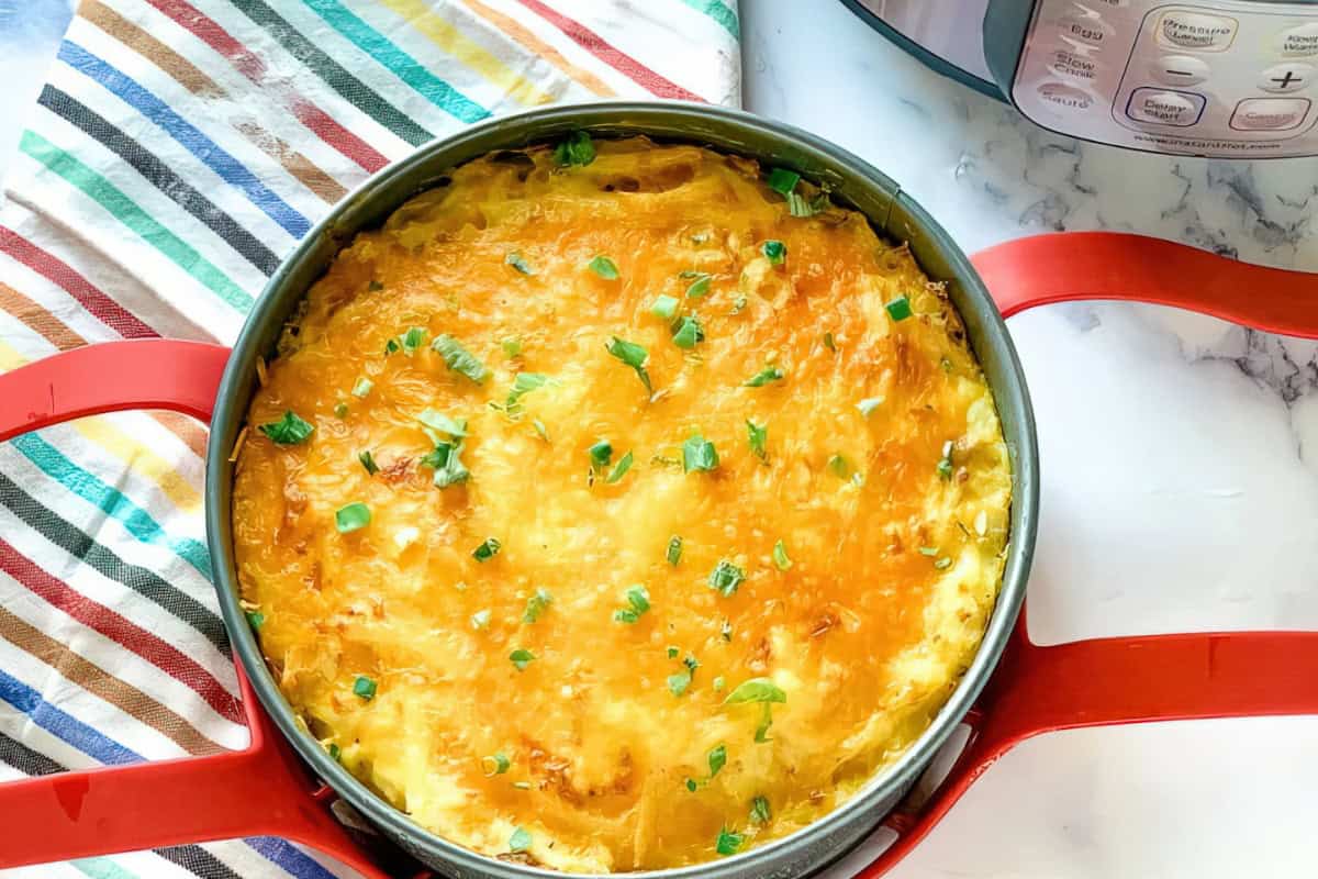 A cheesy baked dish in a round pan with handles, garnished with chopped green herbs. It rests on a colorful striped cloth, next to a kitchen appliance on a marble surface.